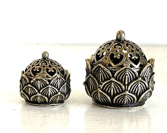 May include: Two gold metal incense burners shaped like lotus flowers. The larger burner has a more intricate design with a round top and a base of overlapping petals. The smaller burner has a similar design but is smaller in size.