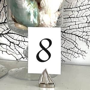 May include: A white card with the number 8 in black is held by a silver boat-shaped place card holder. A glass vase filled with seashells and a decorative coral branch are in the background.