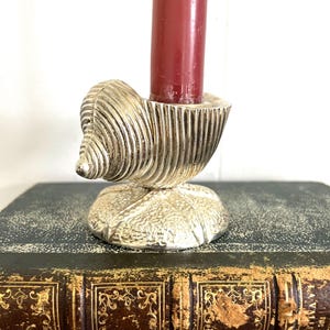May include: A silver-colored seashell-shaped candle holder with a red candle. The candle holder is resting on a dark book with gold accents. The seashell design has detailed ridges.