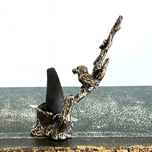 May include: A gold metal incense holder shaped like a tree branch with a bird perched on it. The holder has a cone-shaped incense burner.