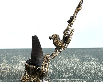 May include: A gold metal incense holder shaped like a tree branch with a bird perched on it. The holder has a cone-shaped incense burner.