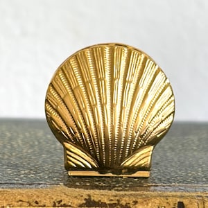May include: A gold-colored, seashell-shaped decorative object. The object has a detailed, textured surface with vertical lines and a scalloped edge. The seashell is set on a dark surface, possibly a book, with gold accents.