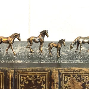 May include: Four miniature bronze horse figurines, each with a different pose, are displayed on a brown leather-bound book.
