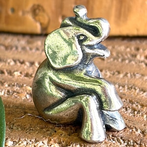 May include: A small, silver-colored elephant figurine in a seated position. The elephant has large ears, a trunk, and detailed legs. The figurine is resting on a textured, brown surface, with a blurred wooden background.