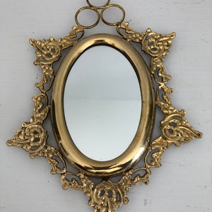 Puede incluir: Un espejo ovalado dorado con un marco ornamentado y decorativo. El marco tiene un dise&ntilde;o floral y tres anillos en la parte superior para colgar.