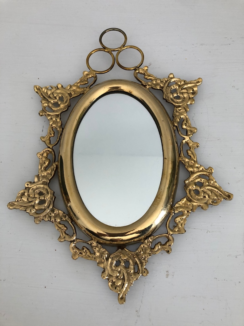 Puede incluir: Un espejo ovalado dorado con un marco ornamentado y decorativo. El marco tiene un dise&ntilde;o floral y tres anillos en la parte superior para colgar.