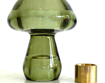 May include: A translucent olive green glass mushroom-shaped object with a wide cap and a cylindrical base, positioned next to a small, plain, cylindrical gold-colored metal ring. The glass piece has a smooth, reflective surface and a hollow interior.