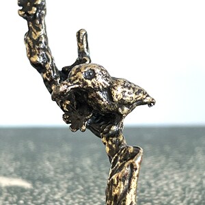 May include: A gold-toned metal bird perched on a branch, the bird is facing right and has a detailed, textured surface.