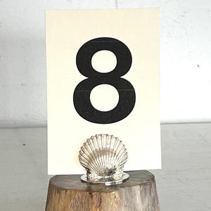 May include: A wooden table number holder with a silver seashell design. The number 8 is displayed on a white card.