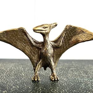 May include: A gold-colored metal figurine of a pterodactyl with its wings outstretched. The figurine is detailed with textured wings and a feathered body.