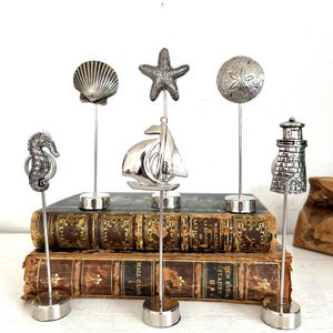 May include: A collection of silver-toned nautical-themed figurines, including a seahorse, seashell, starfish, sailboat, sand dollar, and lighthouse. Each figurine is mounted on a slender metal rod with a circular base. The figurines are displayed on top of antique books.