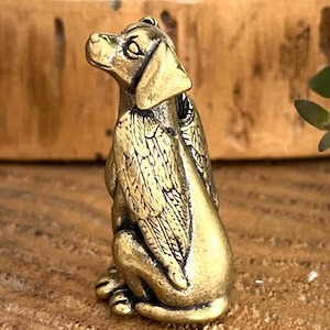 May include: A small, golden-colored figurine of a dog with feathered wings. The dog is in a seated position, with its head turned slightly to the side. The wings are detailed with intricate feather patterns. The figurine is made of metal and has a vintage appearance.