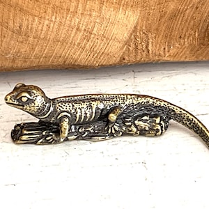 May include: A small, bronze-colored lizard figurine with a detailed, textured surface. The lizard is posed on a base with a leafy design.