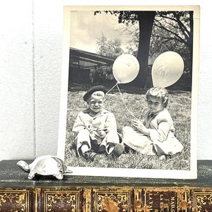 May include: Black and white photograph of two children holding balloons. The balloons are decorated with drawings of elephants. The photo is framed and sitting on a book.
