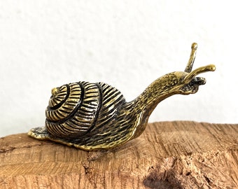 May include: A gold-colored metal snail figurine with a detailed shell and a textured body. The snail is perched on a piece of wood.
