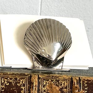May include: Silver seashell shaped desk organizer for holding notes or cards. The organizer is sitting on a brown leather bound book.