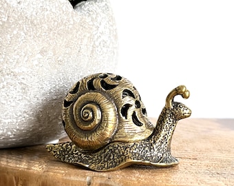 May include: A gold-colored metal snail figurine with a detailed, intricate design. The snail's shell is openwork and the body is textured. The figurine is sitting on a wooden surface.