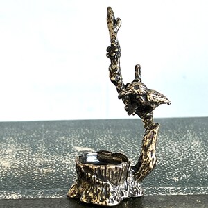 May include: A silver-toned metal ring holder shaped like a tree stump with a bird perched on a branch. The bird is facing to the right and has a detailed design.