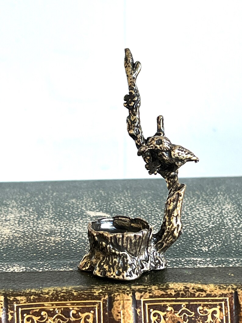 May include: A silver-toned metal ring holder shaped like a tree stump with a bird perched on a branch. The bird is facing to the right and has a detailed design.