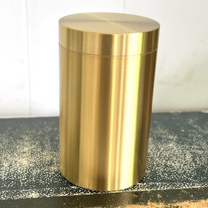 May include: A cylindrical, gold-colored container with a matching lid. The container has a smooth, metallic finish and a simple, modern design. The container is approximately 6 inches tall and 3 inches in diameter.