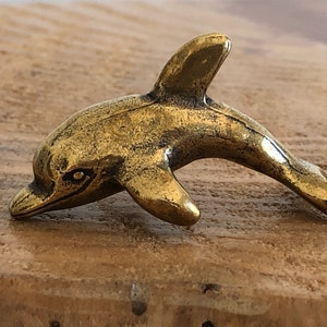 May include: A small, gold-colored dolphin figurine. The dolphin is in a leaping pose with its head tilted down.