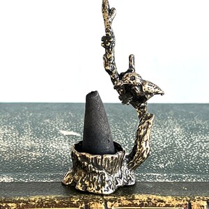 May include: A small, metal incense holder shaped like a tree stump with a bird perched on a branch. The holder is designed to hold a cone incense.