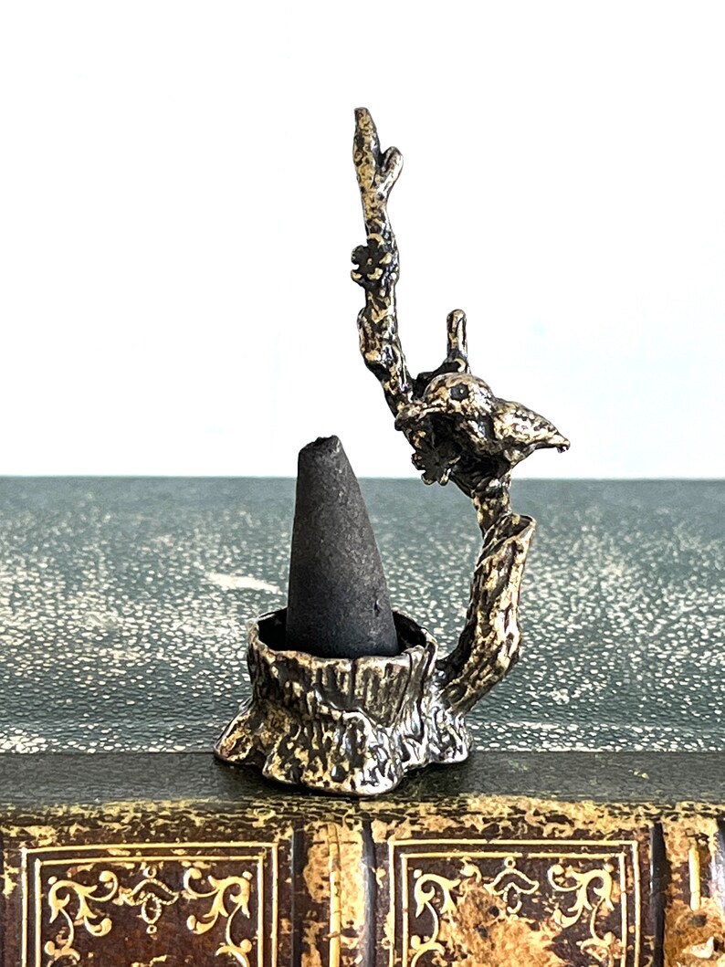 May include: A small, metal incense holder shaped like a tree stump with a bird perched on a branch. The holder is designed to hold a cone incense.