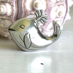 May include: A small, silver-colored whale figurine with a stylized design. The whale has a curved body, a smiling face, and a decorative spout. The background features a textured, light purple object.