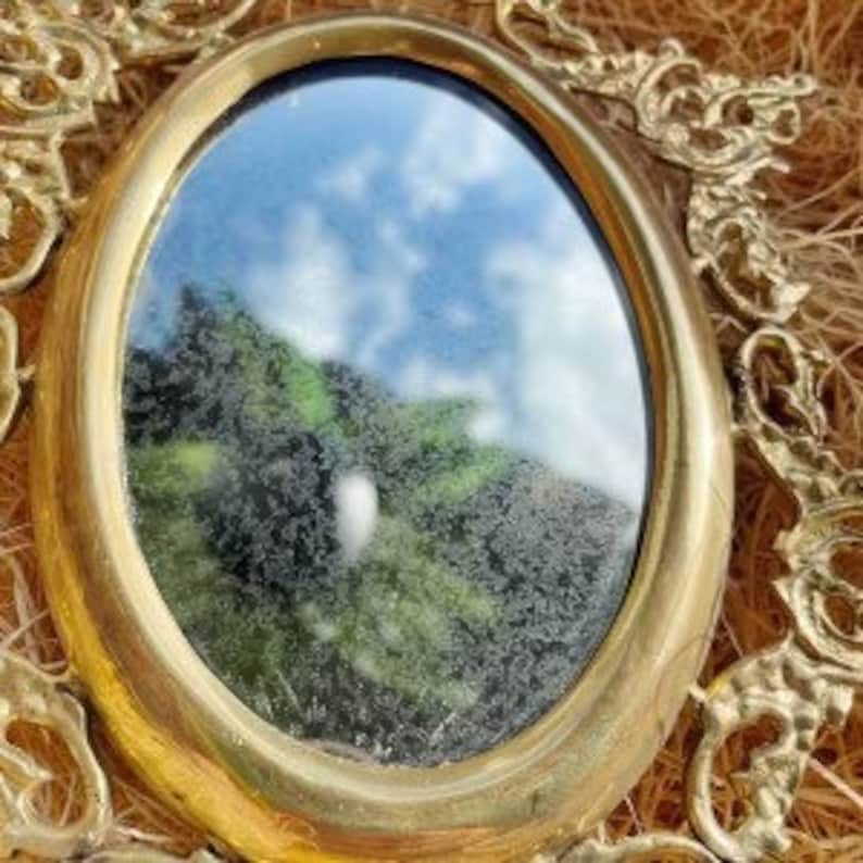 Puede incluir: Un peque&ntilde;o espejo ovalado con marco de lat&oacute;n que refleja un cielo azul con nubes blancas y follaje verde.