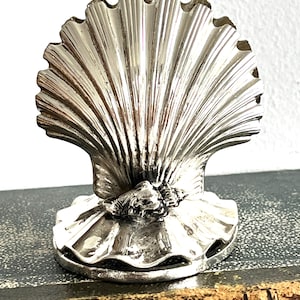 May include: A silver-colored seashell-shaped object with a detailed, ridged design. The object has a base and a detailed, textured design. The seashell is sitting on a dark surface.