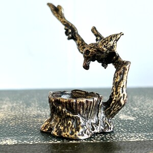 May include: A small, bronze sculpture of a bird perched on a tree stump. The bird is facing to the right and has its wings spread out. The tree stump is detailed with bark and has a small, round opening in the top.