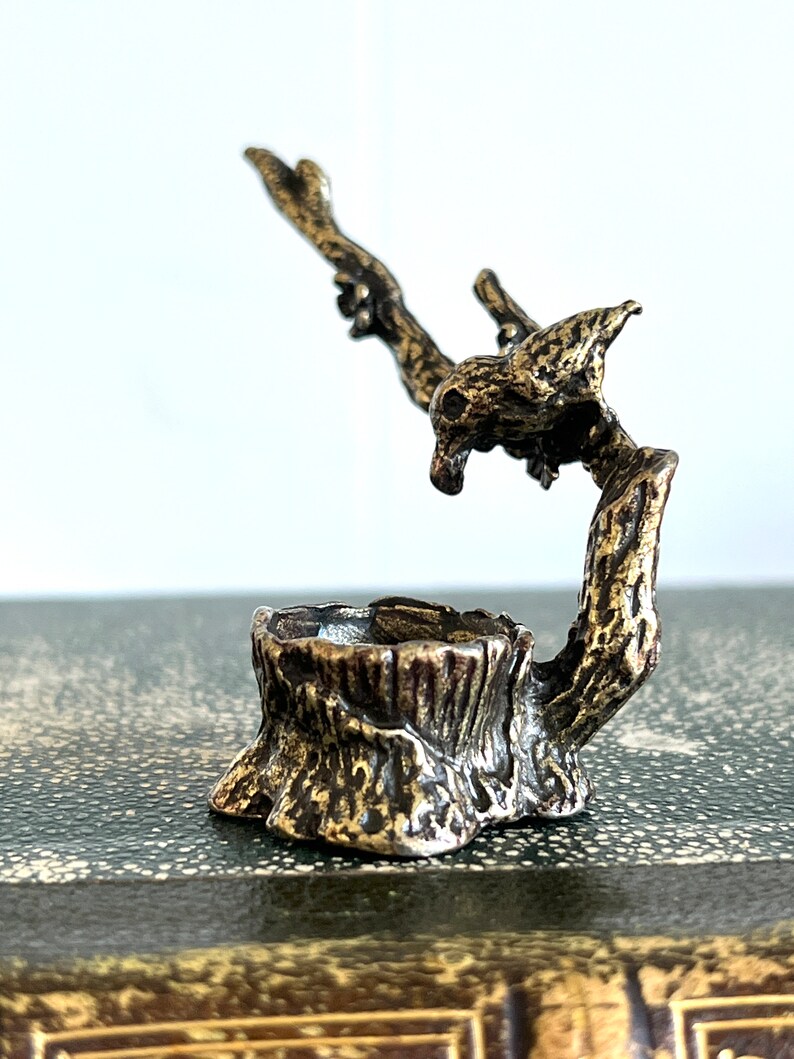 May include: A small, bronze sculpture of a bird perched on a tree stump. The bird is facing to the right and has its wings spread out. The tree stump is detailed with bark and has a small, round opening in the top.