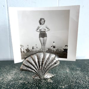 May include: A silver seashell photo holder with a black and white photo of a woman in a swimsuit standing on a beach.