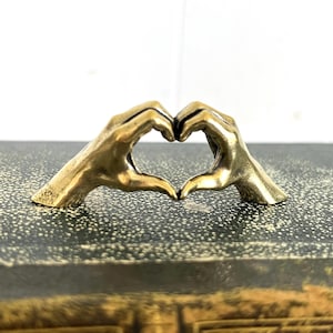 May include: A golden-colored sculpture of two hands forming a heart shape. The hands are detailed and appear to be made of metal. The sculpture is placed on a textured, dark surface.