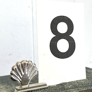 May include: A silver seashell-shaped place card holder next to a white card with a large black number 8 printed on it. The card is resting on a dark, textured surface.