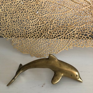 May include: A small, gold-colored brass dolphin figurine. The dolphin is in a leaping pose with its tail raised and its head tilted forward.