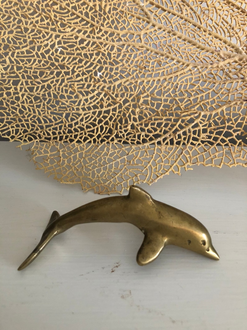 May include: A small, gold-colored brass dolphin figurine. The dolphin is in a leaping pose with its tail raised and its head tilted forward.