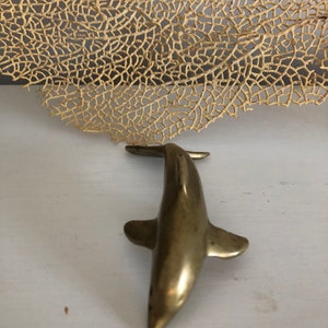 May include: A small, gold-colored metal dolphin figurine. The dolphin is facing forward with its tail up and its fins out.