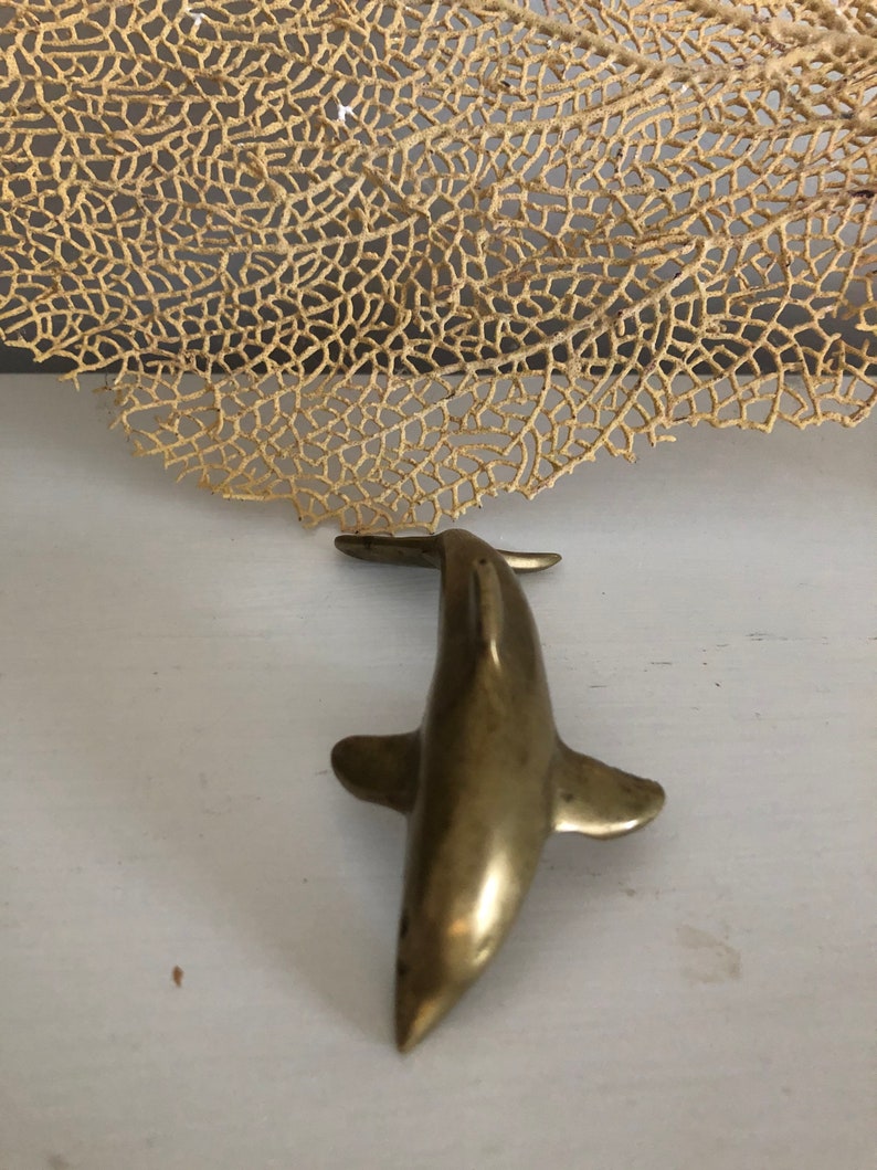 May include: A small, gold-colored metal dolphin figurine. The dolphin is facing forward with its tail up and its fins out.