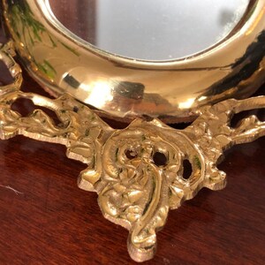 Puede incluir: Un peque&ntilde;o espejo redondo, adornado, de color dorado, con un marco decorativo. El marco tiene un dise&ntilde;o floral y est&aacute; hecho de metal.