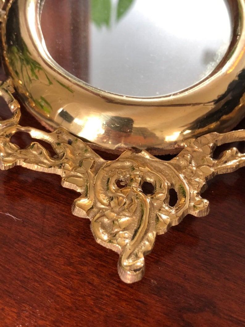 Puede incluir: Un peque&ntilde;o espejo redondo, adornado, de color dorado, con un marco decorativo. El marco tiene un dise&ntilde;o floral y est&aacute; hecho de metal.