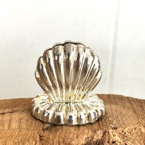 May include: Silver seashell shaped card holder. The holder is detailed with a textured shell design.