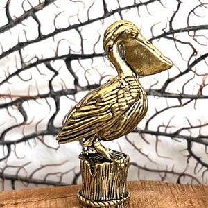 May include: A golden pelican figurine perched on a wooden stump base. The pelican has detailed feather and beak features. The base is intricately designed with a rope-like border. The background features a white and brown branch-like pattern.