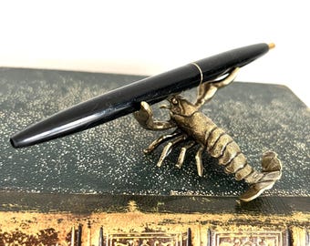 May include: A black pen resting in a gold-toned lobster-shaped pen holder. The pen holder is sitting on top of a vintage book with a gold-embossed design.