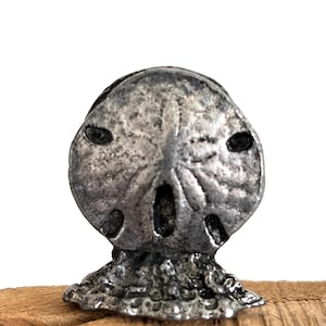 May include: A silver-colored sand dollar sculpture with detailed texture and dark eye-like features. The sculpture is mounted on a piece of natural wood, showcasing a coastal or nautical theme.