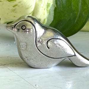 May include: A small, silver-colored bird figurine with etched details. The bird has a rounded body, a small beak, and a curved line detail on its side. The figurine is sitting on a white surface, with green and white foliage in the background.