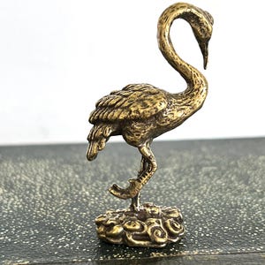 May include: A small, detailed brass figurine of a crane. The bird is depicted with its neck curved, standing on a decorative base. The crane's feathers and features are intricately carved, showcasing a vintage aesthetic.