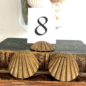 May include: Three gold-colored metal seashell-shaped place card holders with ribbed textures. One holder supports a white rectangular card featuring the black printed number 8. The items are arranged on top of two stacked antique books with leather spines.