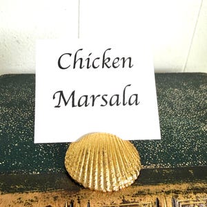 May include: A gold-colored seashell place card holder with the words "Chicken Marsala" printed on a white card. The seashell rests on a vintage book with a dark green cover.