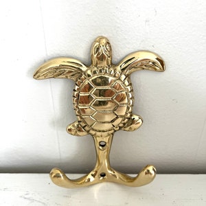 May include: A gold-toned brass sea turtle hook with two hooks on the bottom. The turtle is detailed with a textured shell and has a realistic design.